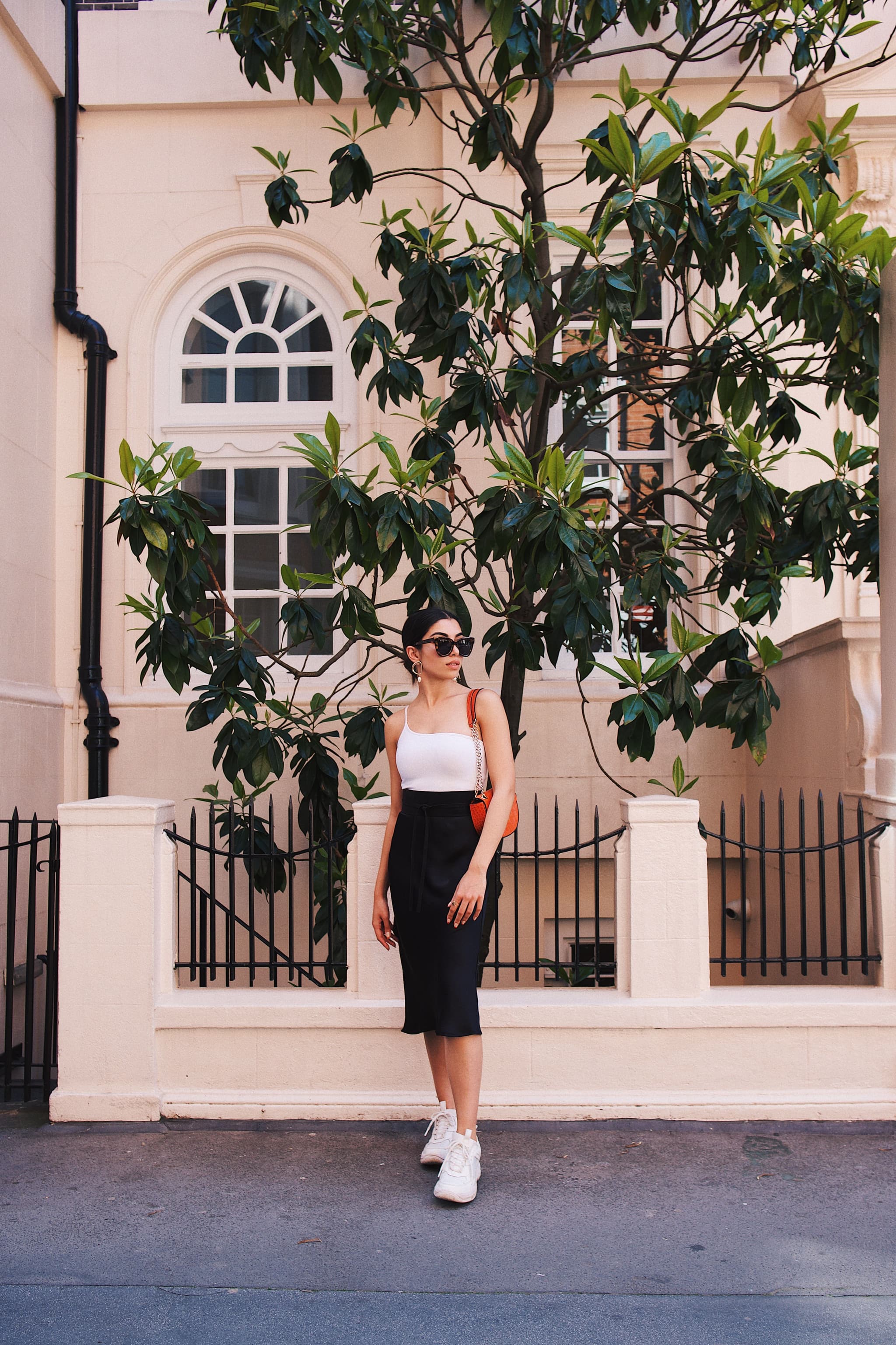 A woman stands on a sidewalk in front of a building with a large leafy tree, embodying the perfect image for selecting the right brand ambassador. She is wearing a white tank top, black skirt, white sneakers, and sunglasses while holding an orange bag.