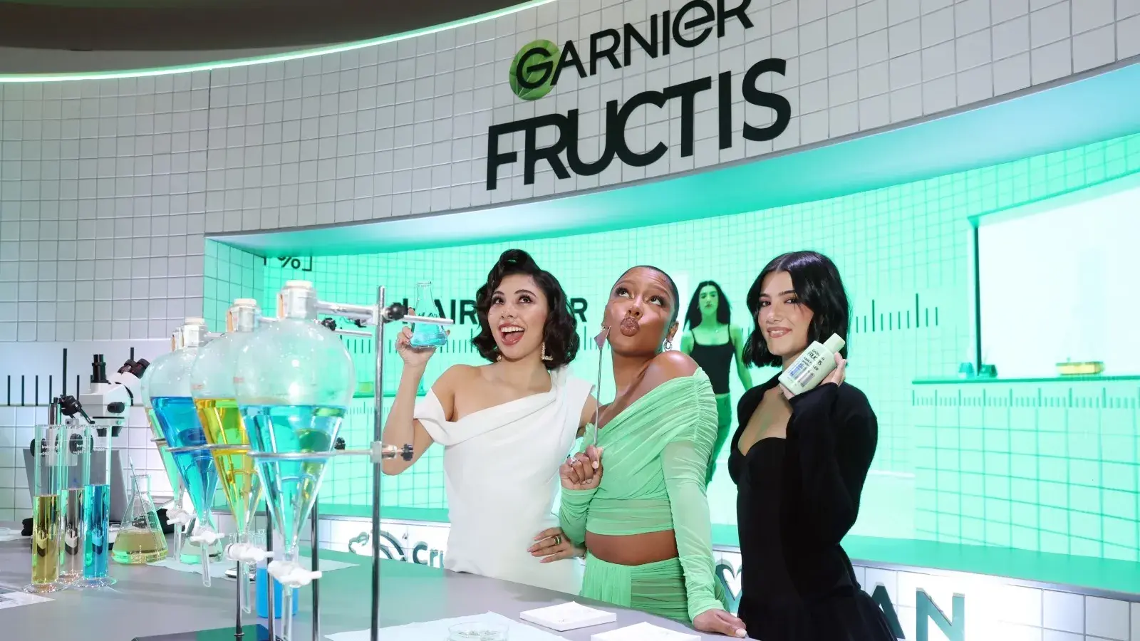 Trois femmes posant avec des produits garnier fructis lors d'un événement promotionnel avec un laboratoire stylisé en arrière-plan.