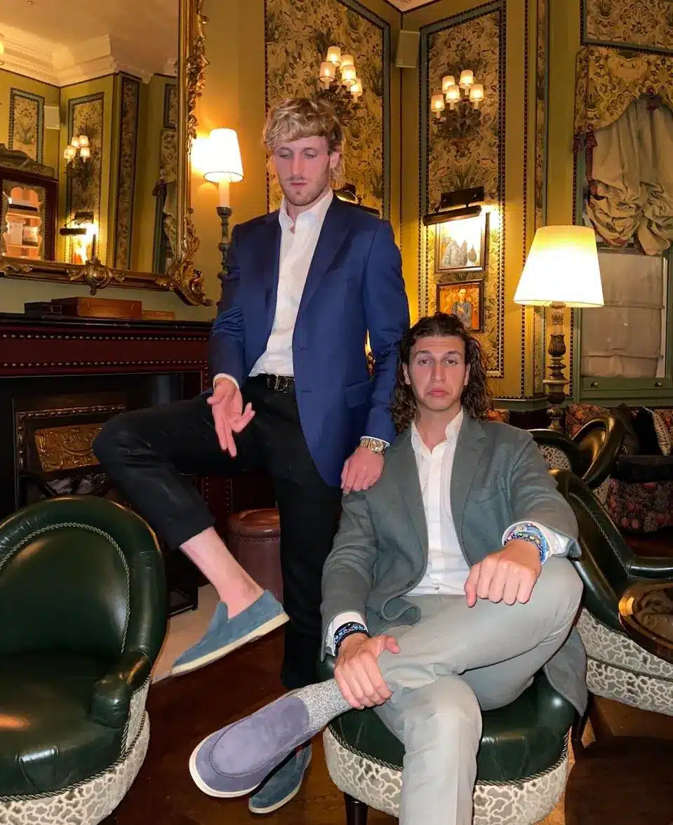 Two men in semi-formal attire pose in a luxurious room with ornate decor, reminiscent of a L’Oreal Paris ad. One stands leaning on a chair, exuding confidence, while the other sits with his legs crossed, embodying elegance and sophistication.