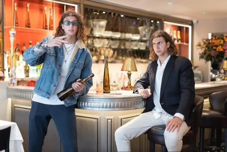 Two men with long hair are posing at a bar. The man on the left, sporting sunglasses and a denim jacket, holds a wine bottle. The man on the right is dressed in a blazer and white pants, making a quirky facial expression. With such distinct styles, they could easily be in an ad for L'Oréal Paris.