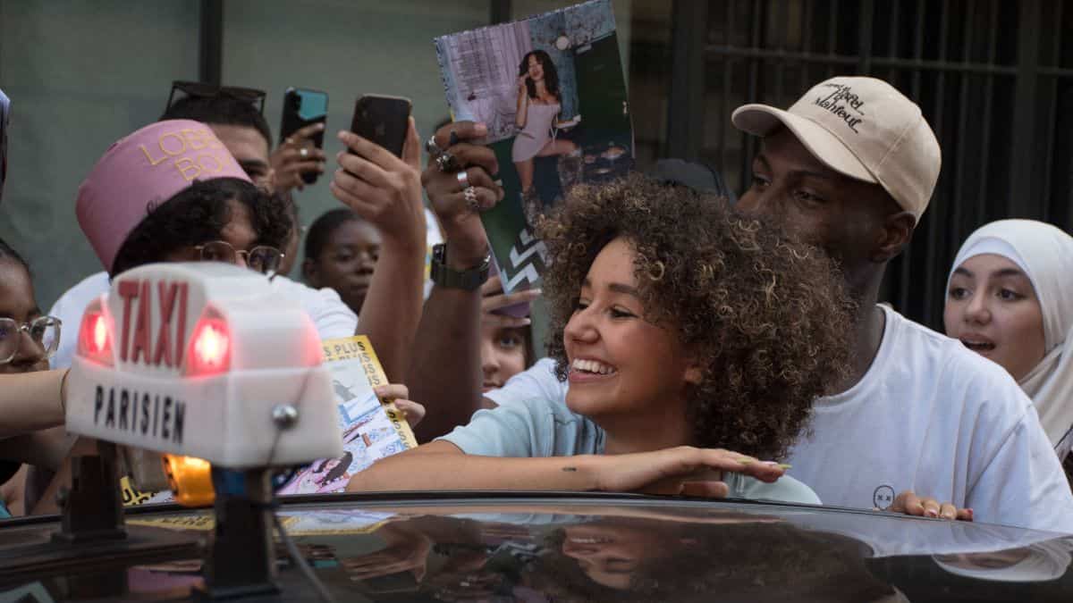 A crowd of people are standing around and behind a taxi, some holding up their phones and magazines for autographs or photos. The taxi sign shows "Parisien.